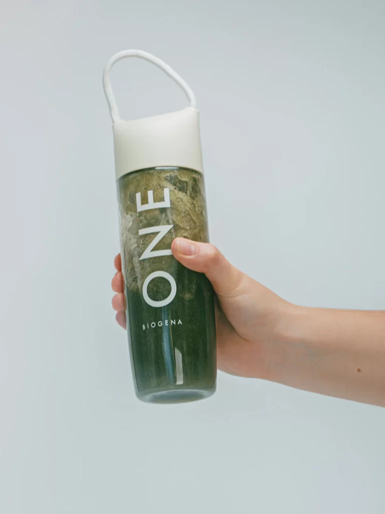A person holding a Biogena One shaker bottle filled with a dark green wellness drink or smoothie against a plain light gray background.