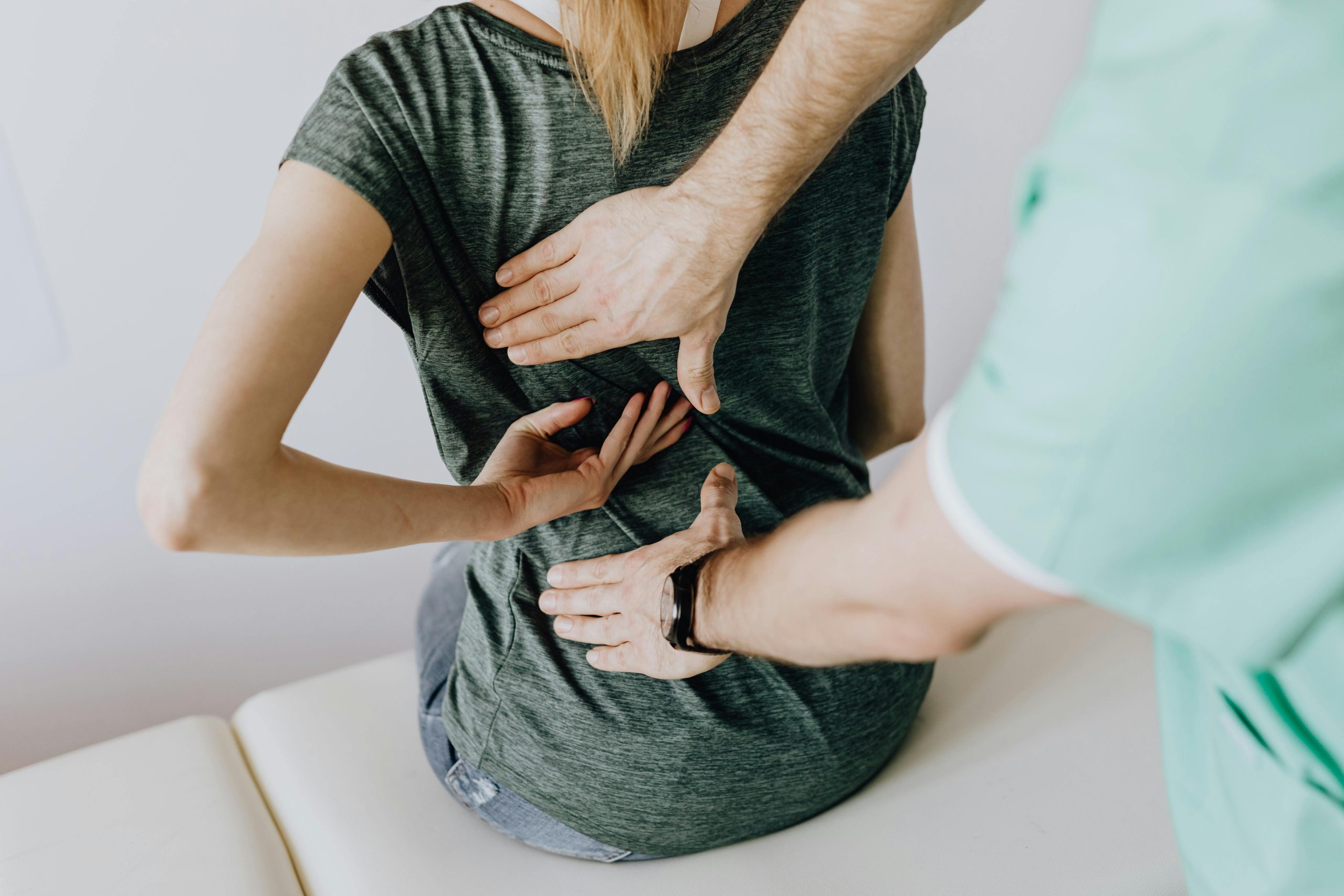 A clinician in green scrubs performing a physical back assessment on a female patient sitting on an examination table, placing hands on her upper and lower back.