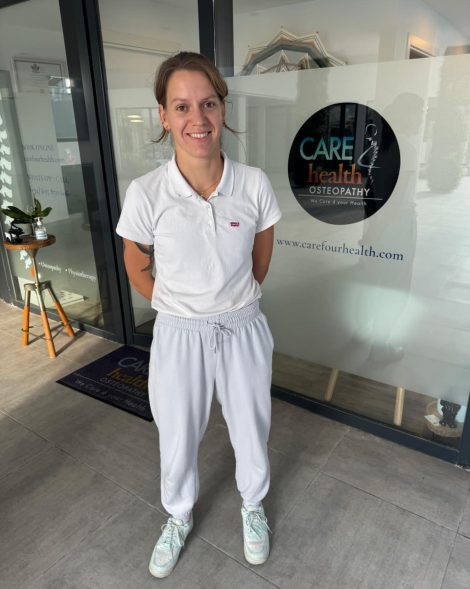 A smiling female practitioner stands inside the bright entryway of Carefour Health Osteopathy clinic, with the company logo and website URL visible on the frosted glass wall behind her.