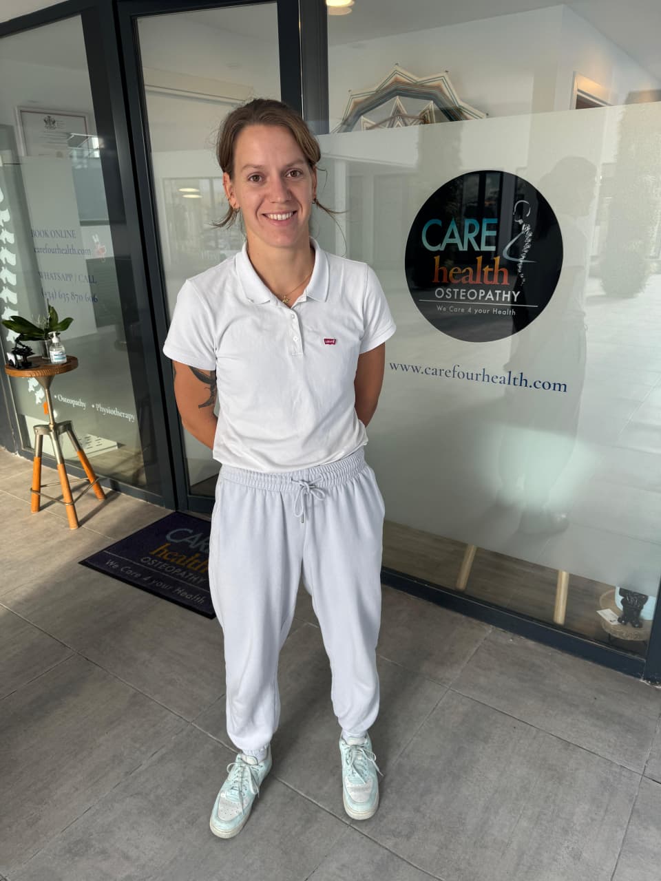 A smiling female practitioner stands inside the bright entryway of Carefour Health Osteopathy clinic, with the company logo and website URL visible on the frosted glass wall behind her.