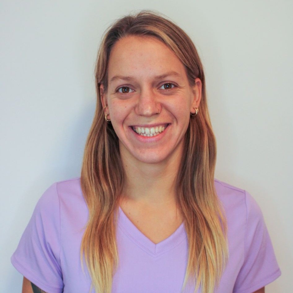 Headshot portrait of a smiling young woman with long blonde-tipped hair wearing light purple v-neck medical scrubs against a plain off-white background.