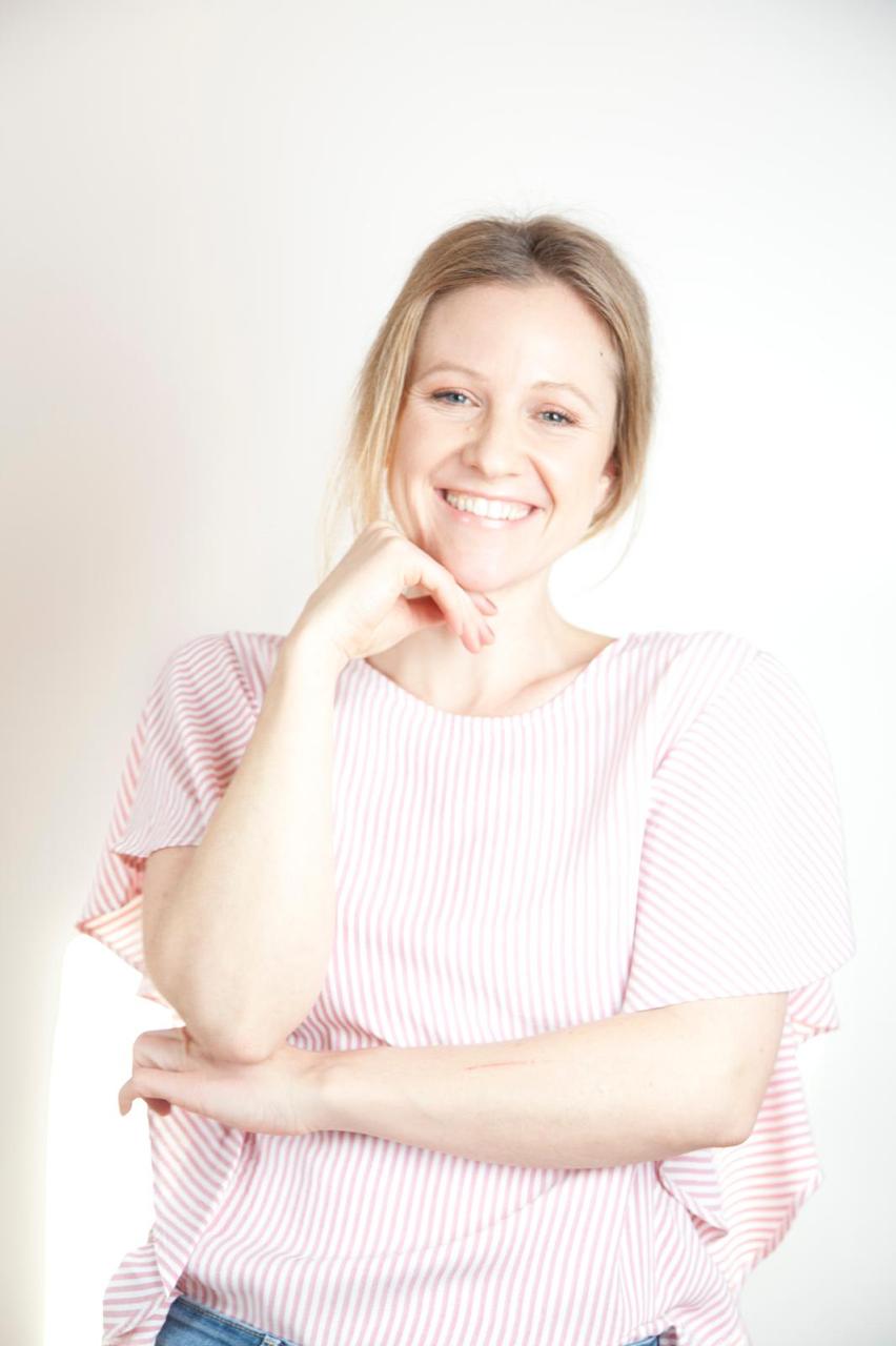 Headshot portrait of a smiling young woman with long blonde-tipped hair wearing light purple v-neck medical scrubs against a plain off-white background.